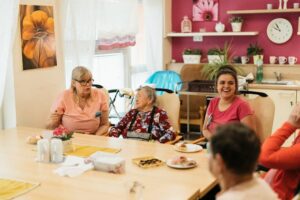 Care staff and elderly residents share a cheerful meal at a sunlit dining room table in a care home.