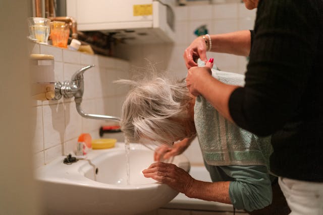 Caregiver rinsing an elderly person's hair over a bathroom sink with a towel draped over their shoulders.