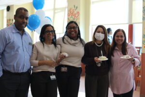 Five colleagues posing together at an indoor celebration, holding cake slices with blue and white balloons in the background.