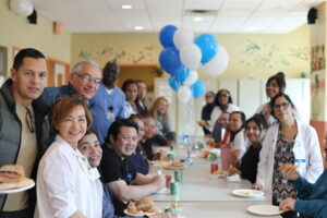 Group of healthcare workers and guests gathered around a long table for a celebration, blue and white balloons in the background, everyone smiling.