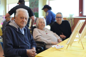 Elderly man in a wheelchair in the foreground, painting at a shared table with others in the background.