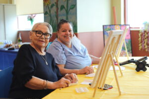 Two smiling women sit at a yellow-draped table with easels, engaged in a craft activity in a bright room.