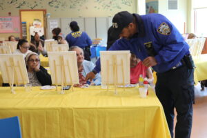NYPD officer in blue uniform assisting elderly women with painting at a table during a community art event.