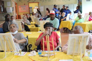 Elderly people painting at easels around yellow tables in a bright room; some wear masks, with staff in blue uniforms in the background.