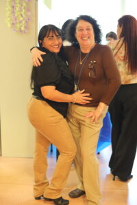 Two women hug and pose for a cheerful photo indoors, smiling at the camera.