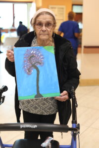 Senior woman with glasses and a pink headband holds a blue painting of a tall, stylized flower in a bright hallway, using a walker nearby.