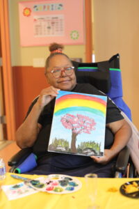 Smiling woman in a wheelchair holding a colorful painting of a pink-blossomed tree under a rainbow sky.