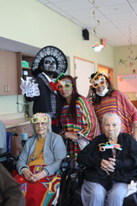Group of people in colorful costumes and masks posing at a party in a care facility, including elderly in wheelchairs and festive decorations overhead.
