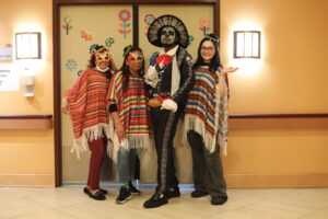 Four people in colorful ponchos posing together in a hallway; one person wears Day of the Dead makeup and a black ornate hat, all smiling at the camera.